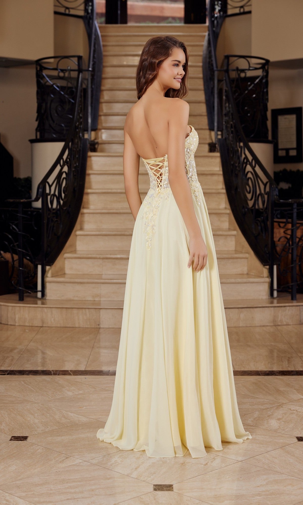 A woman wearing the Nox Anabel C1617 strapless sweetheart prom dress with subtle floral embellishments stands on a marble floor before a staircase, facing away from the camera.